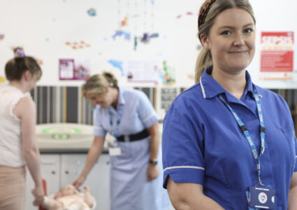 Health Visitor in uniform smiling in front of health visitor colleague, mother and baby in clinic